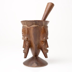 Könnte beinhalten: Ein handgeschnitztes Mörser- und Stößel-Set aus Holz. Der Mörser ist mit drei Gesichtern verziert und hat eine konische Form. Der Stößel ist ein einfacher, abgerundeter Stab. Das Set besteht aus dunkelbraunem Holz und steht auf weißem Hintergrund.