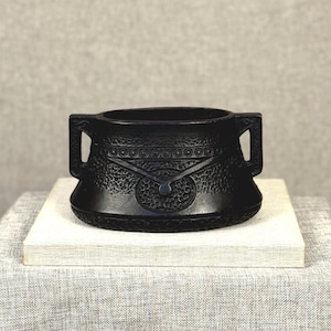 May include: A black, decorative ceramic pot with two handles. The pot has a textured surface with a circular design in the center. It sits on a white rectangular block, placed on a gray linen tablecloth.