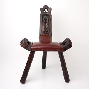 May include: A dark wood chair with a carved back and a red leather seat. The chair has three legs and decorative elements on the arms. The back of the chair features a carved plant design. The chair is a unique piece of furniture.