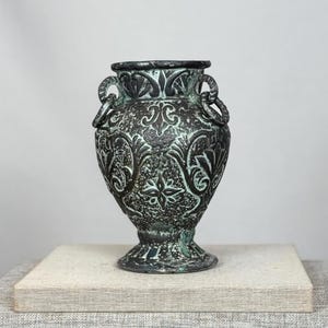May include: A dark, weathered metal vase featuring a textured surface with raised floral and star-shaped patterns. The vessel has a wide rim, a tapered body, and two small ring-shaped handles attached to the sides, finished in a patina of black and oxidised green.
