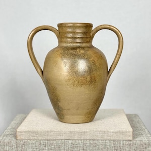 May include: A gold-toned ceramic vase with two arched handles. The vase has a rounded body and a textured surface with dark brown speckles. The top of the vase has horizontal ridges. The vase is displayed on a square, light-coloured platform.
