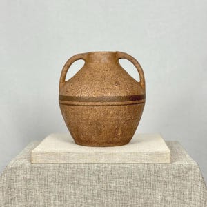 May include: A white ceramic mug with a black handle and the text "But First, Coffee" in a bold, black font. The mug is set against a blurred background. The mug is approximately 10 cm tall.