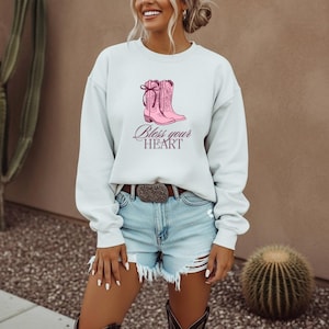 May include: A light gray sweatshirt featuring a pink illustration of cowboy boots with a bow. The words "Bless your HEART" are written in a cursive font below the boots. The model is wearing denim shorts and a belt.