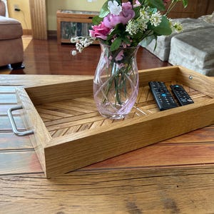 May include: A rectangular wooden serving tray with a geometric pattern, featuring a clear glass vase with pink and white flowers. Two black remote controls rest inside the tray. The tray has metal handles.
