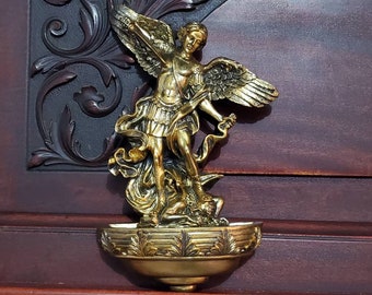 Könnte beinhalten: Ein goldfarbenes Weihwasserbecken zur Wandmontage mit einer detaillierten skulpturalen Darstellung eines Engels, der über einer besiegten Figur steht. Das Becken ist halbkreisförmig mit einem dekorativen Blattmuster am vorderen Rand.