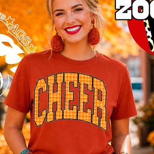 Op de afbeelding: Een roestkleurig t-shirt met korte mouwen met het woord CHEER in een geel en oranje ruitpatroon met een zwarte omlijning. Het ontwerp bevat de tekst ZOOM, PAPETERIE AGS en SUBLIMATION DESIGN PNG FILE in het omliggende gebied.