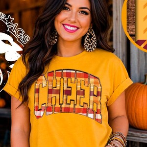 Op de afbeelding: Een mosterdgeel T-shirt met korte mouwen en het woord CHEER in een rood, oranje en geel ruitpatroon op de borst. Het ontwerp heeft een donkere omlijning en een licht gebogen vorm. Het shirt heeft opgerolde mouwen en een ronde halslijn.