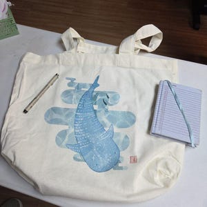 May include: Off-white tote bag featuring a blue whale illustration with a light blue cloud design. The bag has two handles and a small notebook with a blue ribbon. A black pen rests on the bag.