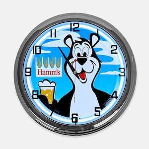 May include: A round wall clock with a silver-colored metal frame. The clock face features a cartoon bear holding a beer, with the Hamm's logo and a blue sky background. The clock hands indicate the time.