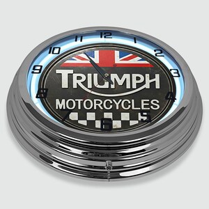 Może przedstawiać: Chromowany zegar ścienny Triumph Motorcycles z niebieskim neonem. Tarcza zegara przedstawia flagę Union Jack i logo Triumph. Zegar ma retro design z szachownicą na dole.