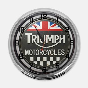 Może przedstawiać: Okrągły zegar ścienny z chromowaną ramką. Tarcza zegara przedstawia logo Triumph Motorcycles, Union Jack i wz&oacute;r szachownicy. Czarne cyfry oznaczają godziny. Wskaz&oacute;wki zegara są czarne i czerwone.