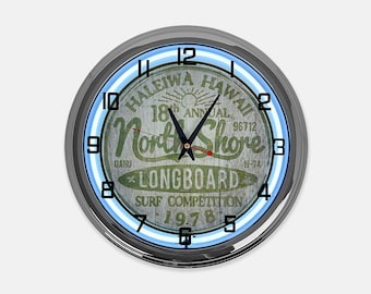 May include: A round wall clock with a silver-colored metal frame and a blue neon light ring. The clock face features a weathered wood-look design with the words "Haleiwa Hawaii North Shore Longboard Surf Competition 1978".