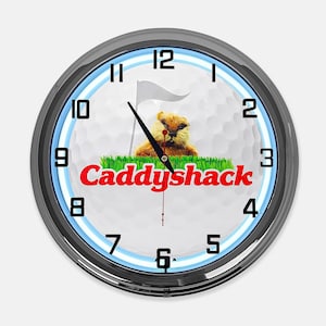 May include: A round wall clock with a chrome frame and a white face featuring a golf ball texture. The center displays a gopher graphic, a golf flag, and the red text Caddyshack. Black hour and minute hands are present with a thin red second hand.