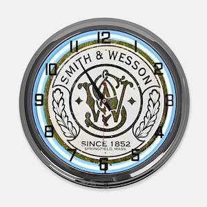 May include: A round wall clock with a black metal frame. The clock face features the "Smith & Wesson" logo in a vintage style, with a light blue and white illuminated ring around the face. The text "SINCE 1852 SPRINGFIELD, MASS." is printed at the bottom.