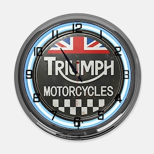 Może przedstawiać: Okrągły zegar Triumph Motorcycles z czarną tarczą, flagą Union Jack i niebieskim neonem. Tarcza zegara zawiera logo Triumph i napis "MOTORCYCLES". Zegar ma chromowaną ramkę.