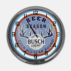 May include: A round wall clock with a silver-colored frame. The clock face is light blue with the words "BEER SEASON" and "BUSCH LIGHT" in blue and white. The clock also has a deer antler design and the words "OPEN ALL YEAR-ROUND".