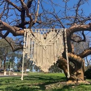 Könnte beinhalten: Ein weißer Makramee-Wandbehang mit geometrischem Muster. Der Behang ist aus natürlichem Baumwollgarn gefertigt und hat einen Fransenrand. Er hängt an einem Ast eines kahlen Baumes.