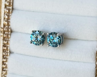 May include: A pair of round, light blue gemstone stud earrings. The earrings are set in a silver metal. The earrings are displayed in a white jewelry box, inside a gold ornate frame. The frame has a decorative design.