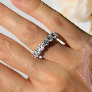 Op de afbeelding: Een zilveren ring met meerdere heldere stenen wordt getoond op een hand met lange, verzorgde nagels. De ring, met zijn sprankelende ontwerp, is de focus. De afbeelding heeft een zachte, elegante uitstraling, met een vage bloemenachtergrond en de merknaam "OZCAL" in de linkerbenedenhoek.