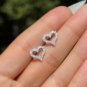 Puede incluir: Un par de pendientes de botón en forma de corazón. Cada pendiente está delineado con pequeñas piedras blancas brillantes. Los pendientes se muestran en una mano, con un fondo verde borroso.
