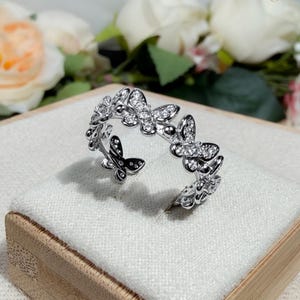 May include: A silver ring with a butterfly design. The ring features multiple butterflies with clear stone accents. It is displayed on a white surface inside a wooden box. The brand name "OZCAL" is visible in the top right corner.