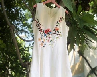 May include: A white sleeveless dress with a floral embroidery design on the front. The embroidery features a branch with pink, red, and blue flowers.