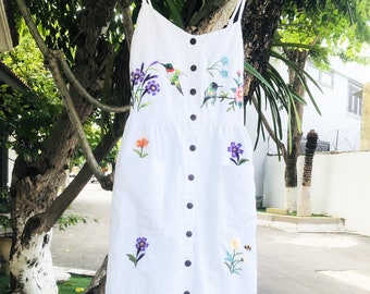 May include: White linen dress with embroidered flowers and hummingbirds. The dress has a button-down front and a relaxed fit.