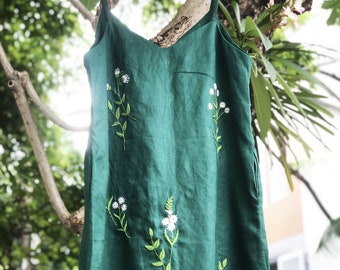 May include: A dark green linen dress with a V-neck and spaghetti straps, adorned with embroidered white flowers and green leaves. The dress is hanging on a pink hanger, showcasing its simple yet elegant design.