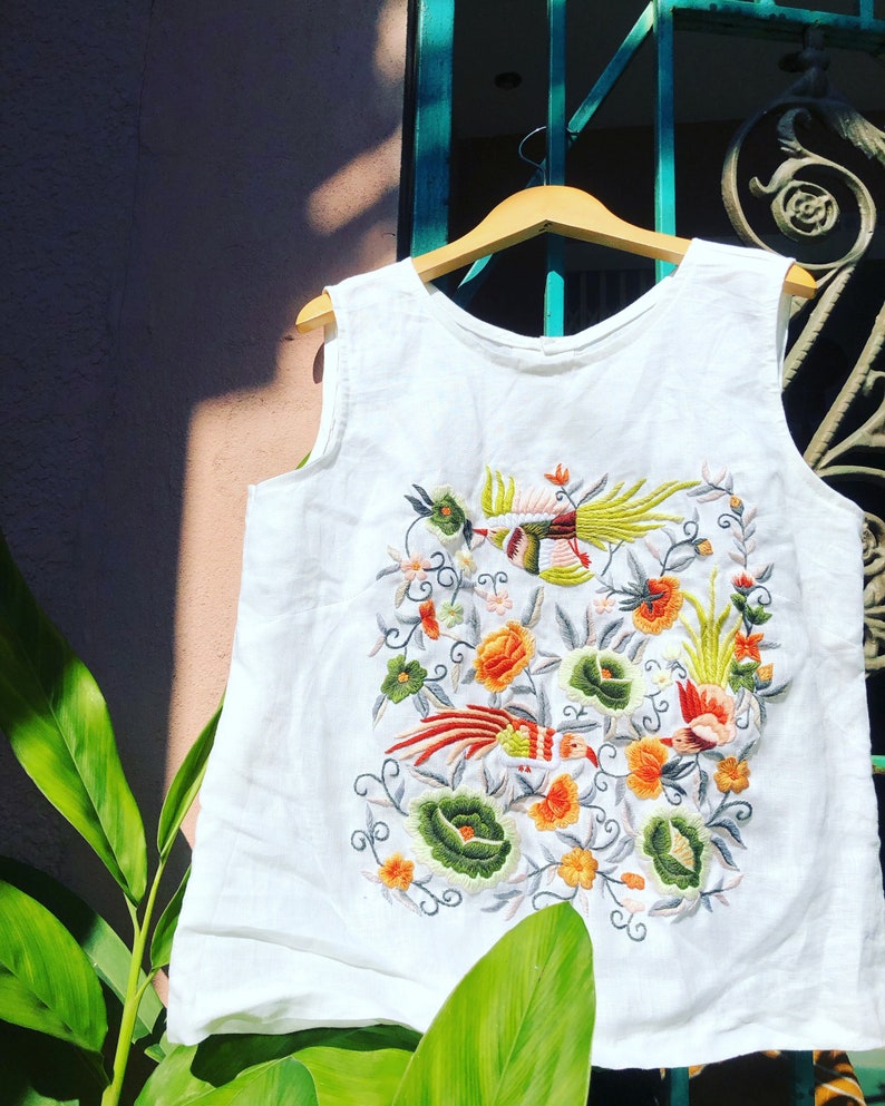 K&ouml;nnte beinhalten: Wei&szlig;es &auml;rmelloses Oberteil mit bunten gestickten V&ouml;geln und Blumen. Die Stickerei zeigt eine Vielzahl von Farben, darunter Rot, Orange, Gelb, Gr&uuml;n und Blau.