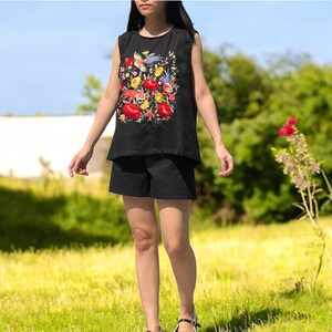 K&ouml;nnte beinhalten: Ein schwarzes &auml;rmelloses Oberteil mit einem floralen Stickmuster mit roten, gelben und blauen Blumen und V&ouml;geln. Das Oberteil wird mit schwarzen Shorts kombiniert.