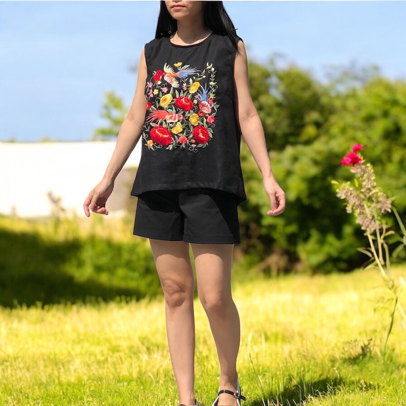 K&ouml;nnte beinhalten: Ein schwarzes &auml;rmelloses Oberteil mit einem floralen Stickmuster mit roten, gelben und blauen Blumen und V&ouml;geln. Das Oberteil wird mit schwarzen Shorts kombiniert.