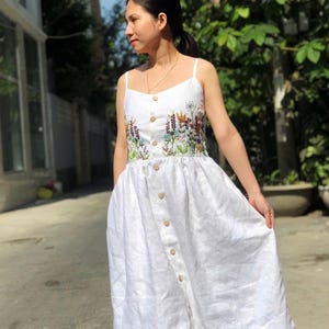 May include: A white linen dress with a floral embroidery design on the bodice. The dress has a button-down front and a flowy skirt.