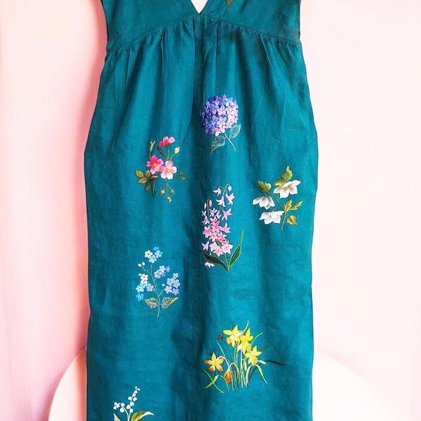May include: Teal-colored dress with a V-neck and cap sleeves. The dress is adorned with embroidered flowers in various colors, including pink, white, and yellow. A gold hanger holds the dress.