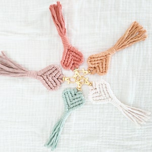 Pode incluir: Seis chaveiros de macram&eacute; em cores pastel, incluindo rosa, p&ecirc;ssego e verde menta, dispostos em forma de estrela. Cada chaveiro apresenta um design de cora&ccedil;&atilde;o tecido e uma borla, conectados a um fecho dourado.
