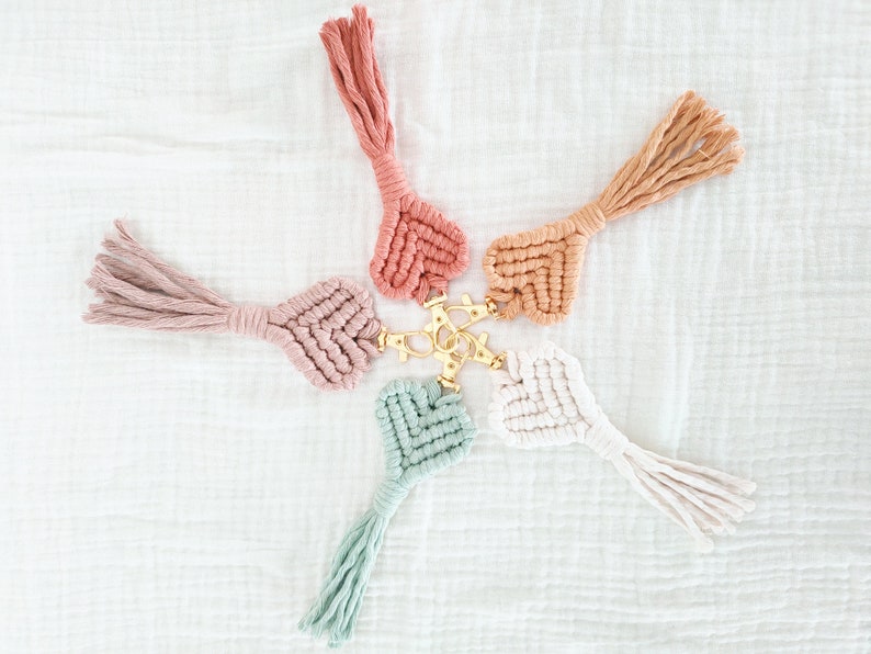 Pode incluir: Seis chaveiros de macram&eacute; em cores pastel, incluindo rosa, p&ecirc;ssego e verde menta, dispostos em forma de estrela. Cada chaveiro apresenta um design de cora&ccedil;&atilde;o tecido e uma borla, conectados a um fecho dourado.