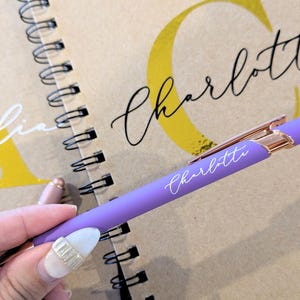 May include: A lilac pen with rose gold detailing and the name "Charlotte" in white script. The pen is placed on a spiral-bound notebook with the name "Charlotte" in large script and the name "Amelia" in white script.