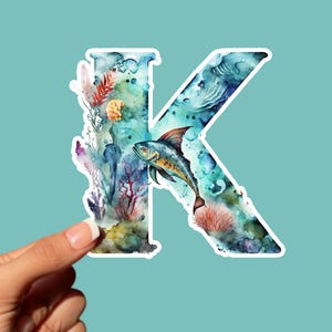 May include: A watercolor sticker of the letter "K" featuring an underwater scene. The letter is filled with a blue and green ocean design, a fish, and coral. The sticker is held by a hand against a teal background.