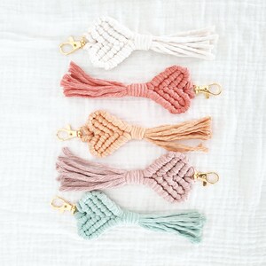 Pode incluir: Cinco chaveiros de macram&eacute; em v&aacute;rias cores: branco, coral, p&ecirc;ssego, malva e verde-azulado. Cada chaveiro apresenta um design de n&oacute; em forma de cora&ccedil;&atilde;o, um fecho dourado e uma borla. Est&atilde;o dispostos sobre uma superf&iacute;cie branca texturizada.