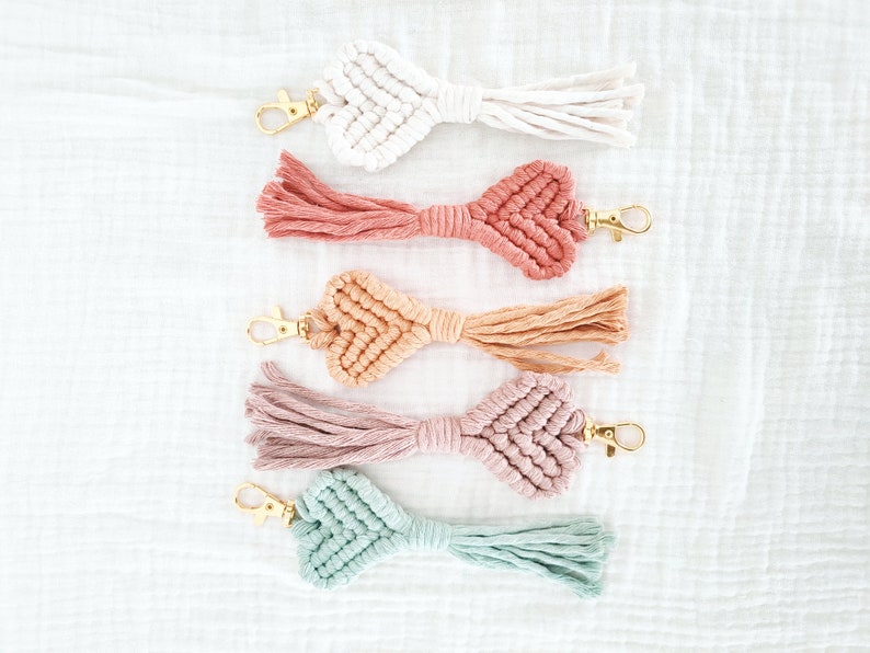 Pode incluir: Cinco chaveiros de macram&eacute; em v&aacute;rias cores: branco, coral, p&ecirc;ssego, malva e verde-azulado. Cada chaveiro apresenta um design de n&oacute; em forma de cora&ccedil;&atilde;o, um fecho dourado e uma borla. Est&atilde;o dispostos sobre uma superf&iacute;cie branca texturizada.