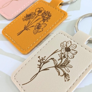 Könnte beinhalten: Drei Leder-Schlüsselanhänger mit Blumenmotiven. Die Schlüsselanhänger sind in den Farben Rosa, Gelb und Creme gehalten. Jeder Schlüsselanhänger zeigt ein anderes Blumenmotiv.