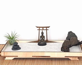 May include: A miniature zen garden with a wooden tray, white sand, and decorative elements. Features a small Buddha statue, a wooden torii gate, rocks, a small plant, and a rake for creating patterns in the sand. The tray is approximately 30 cm long.
