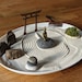 May include: A white ceramic tray with a sand garden. The tray has a small wooden torii gate, a golden buddha statue, a rake, a small pile of stones, and a small patch of green moss.