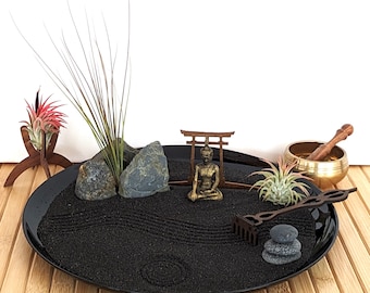 May include: A black sand zen garden with a small wooden torii gate, a miniature Buddha statue, and a rake. There are also small rocks and two air plants.