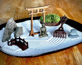 May include: A miniature Zen garden in a black tray. It features a wooden torii gate, a bridge, rocks, a seated Buddha statue, a rake, and a small mossy plant. The sand is raked with patterns, and there are stacked stones.
