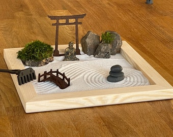 Op de afbeelding: Een houten zen-tuintray met wit zand, een kleine houten brug, een miniatuur torii-poort, een Boeddhabeeld en een stapel zwarte stenen.