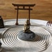 May include: A miniature zen garden with a white sand base, a wooden torii gate, a small golden buddha statue sitting on a gray stone, a rake, and a small patch of green moss.