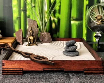 Op de afbeelding: Een houten zen-tuinbak met een zandbasis, een klein Boeddhabeeldje en een stapel grijze stenen. De bak heeft een hark en een houten frame.