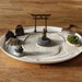 May include: A white circular tray with a sand zen garden. The tray has a wooden torii gate, a small golden buddha statue, a rake, and a small patch of green moss. The sand is raked into a circular pattern.