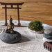 May include: A miniature zen garden with a wooden torii gate, a small golden buddha statue sitting on a gray rock, a small patch of green moss, and a stack of three gray rocks.