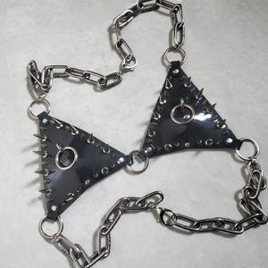 May include: A black, triangular-shaped body harness with silver metal spikes and chain detailing. The harness features two triangle cups connected by a chain and ring hardware. The spikes are evenly spaced around the edges of the triangles.